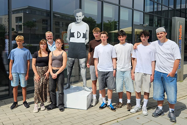 Foto: Unsere Schülerinnen und Schüler mit Lehrer Florian Mersch vor der Erinnerungsfigur für Eric Schildkraut vor der Volksbankhalle auf dem Campus.